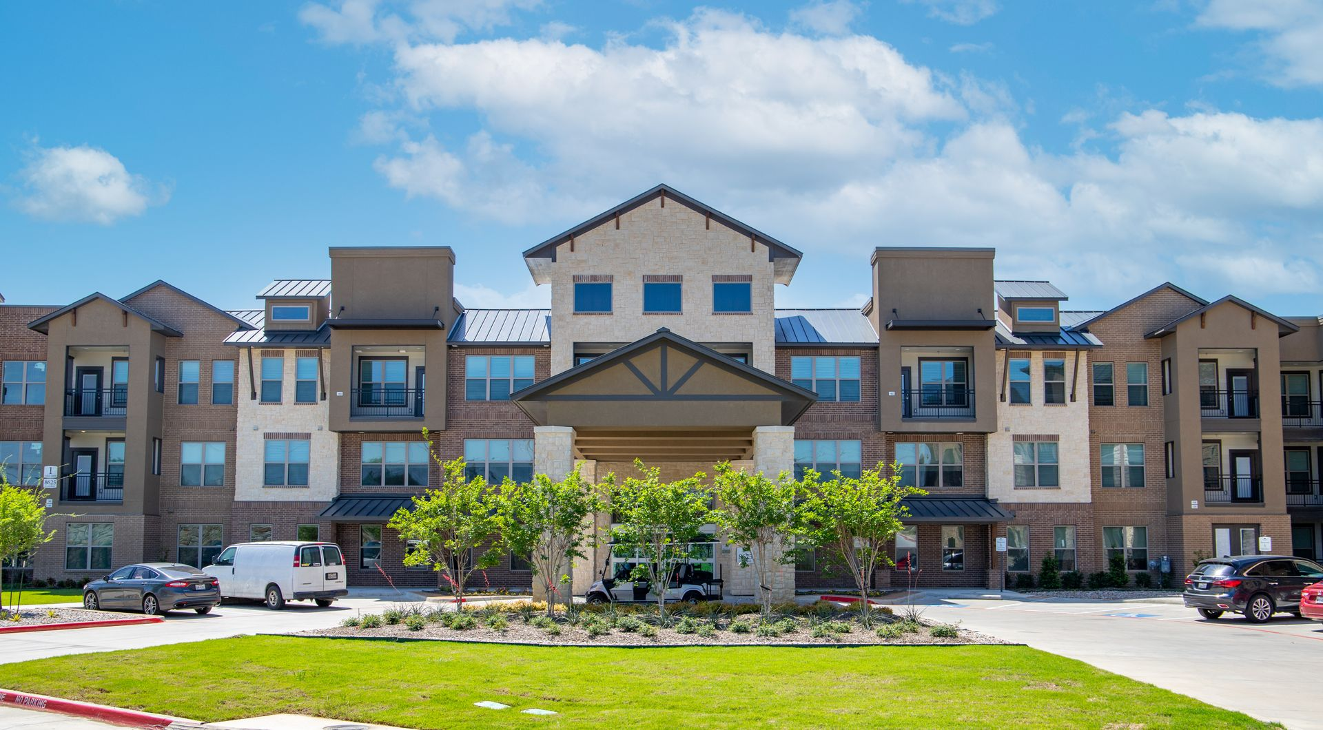 A three-story apartment complex with a beige facade, brown accents, and a central entrance, viewed under a blue sky.