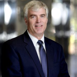 Man in a navy suit and tie smiles in a blurred office setting.