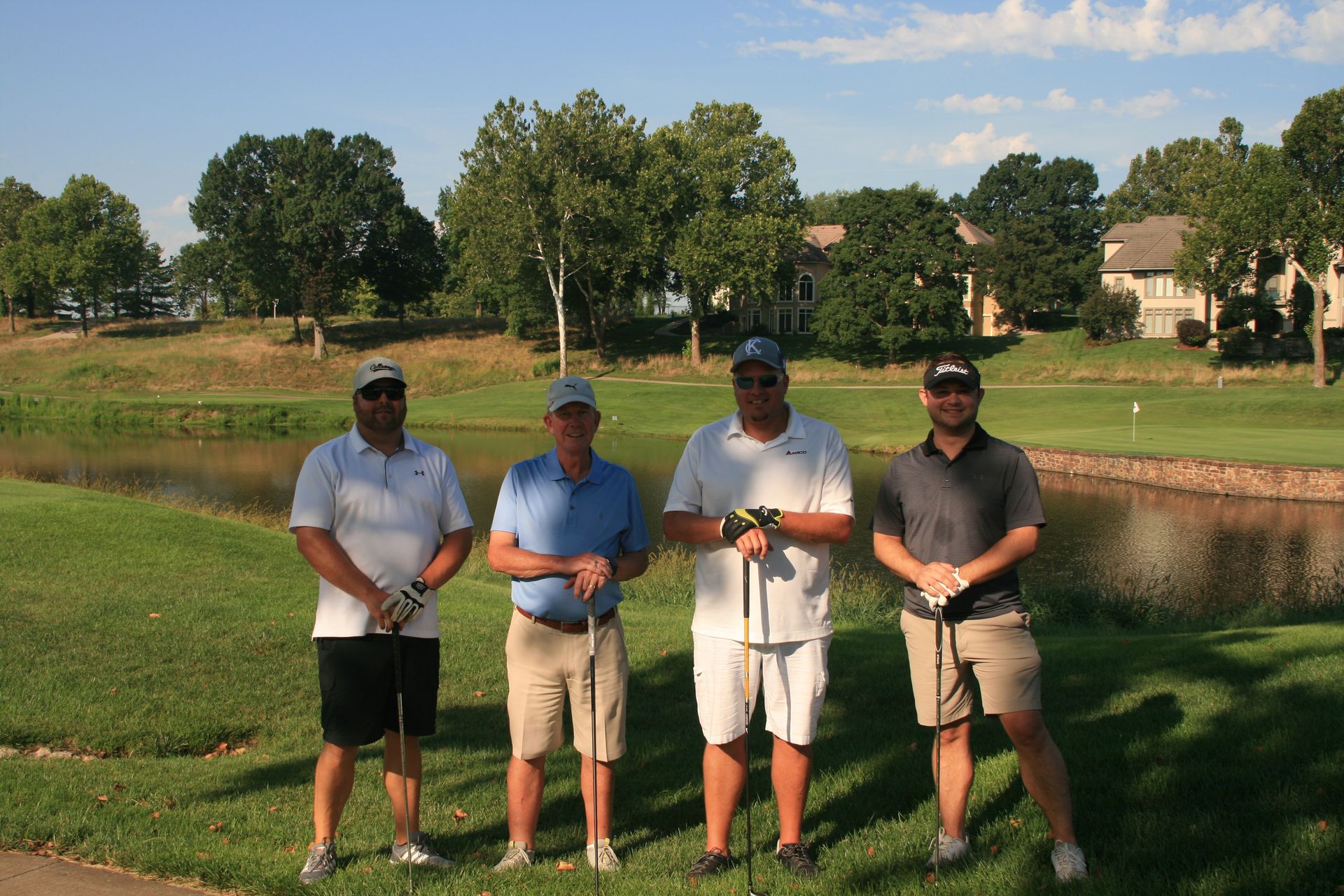 Four men on a golf course, posing with golf clubs. They are standing near a pond on a sunny day.