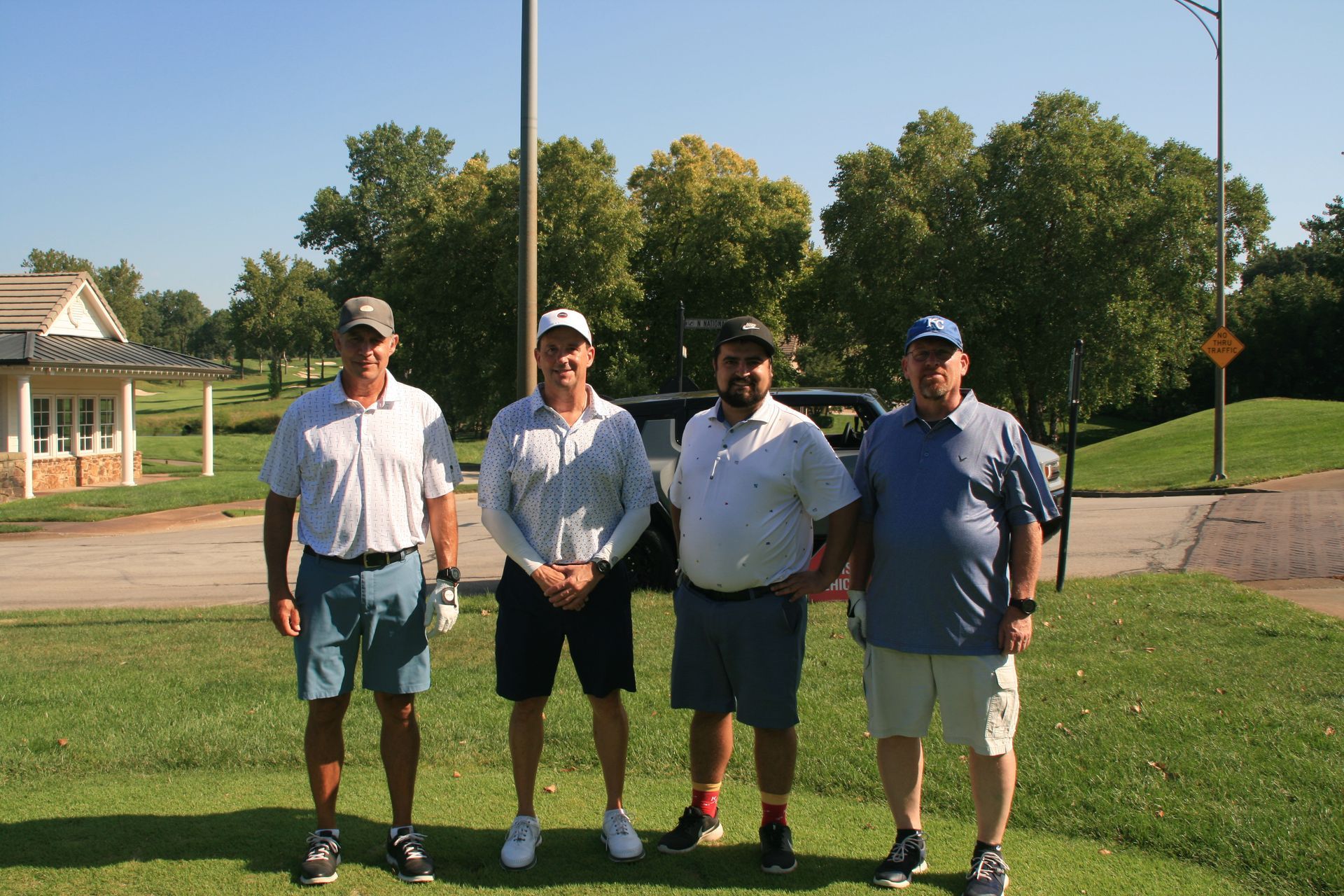 Four men in golf attire pose outdoors on a sunny day; trees in the background.