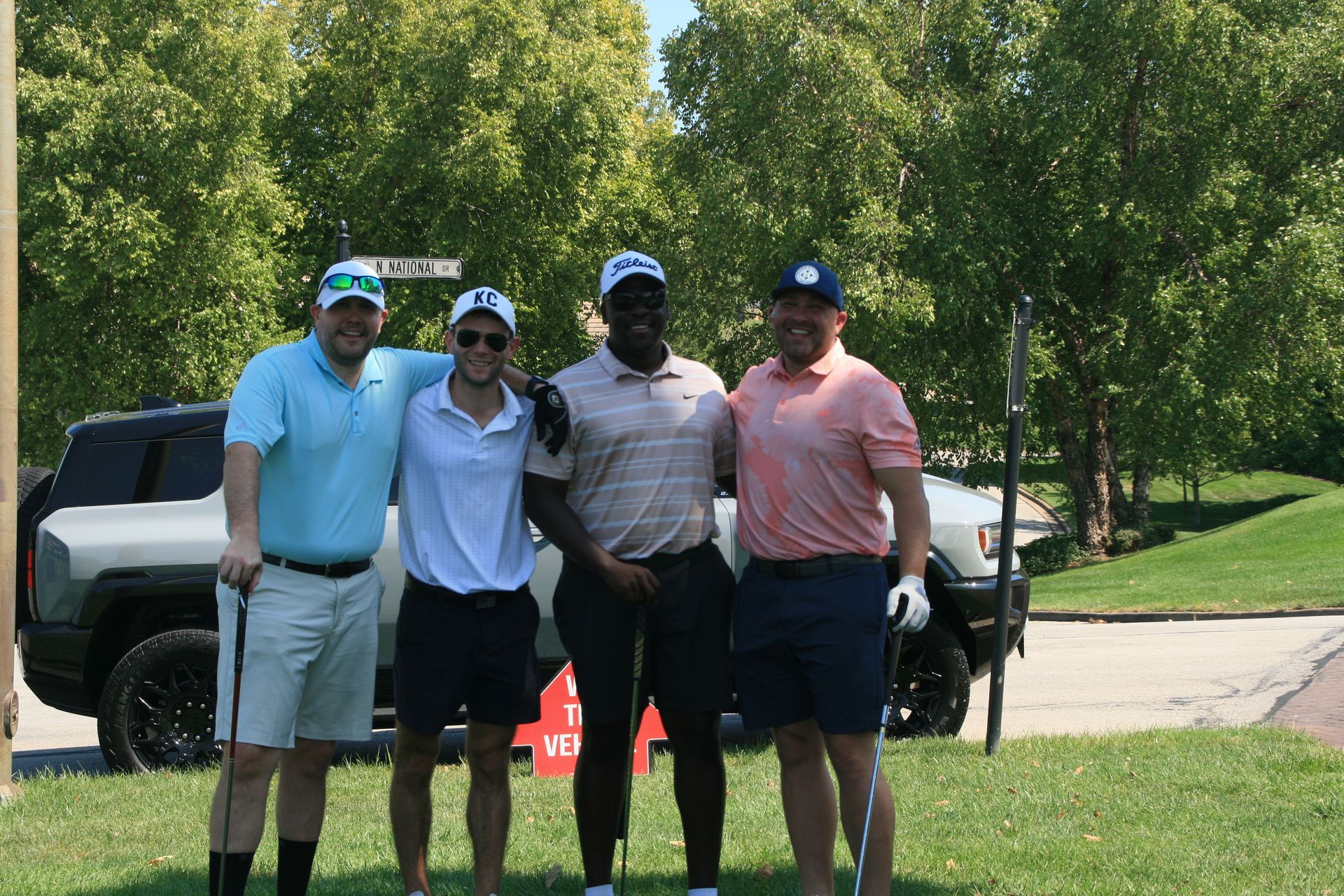 Four men in golf attire pose for a photo outdoors. They stand on grass near a car and trees.