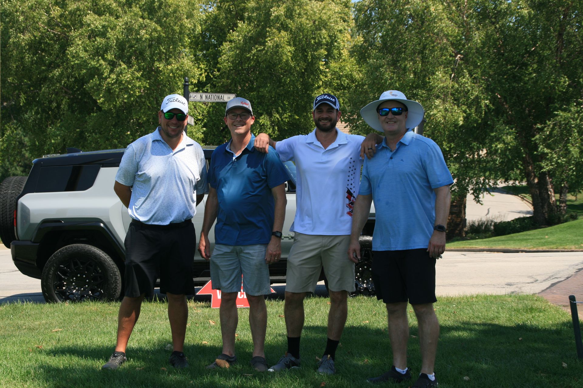 Four men in golf attire standing near a white SUV on a sunny day.
