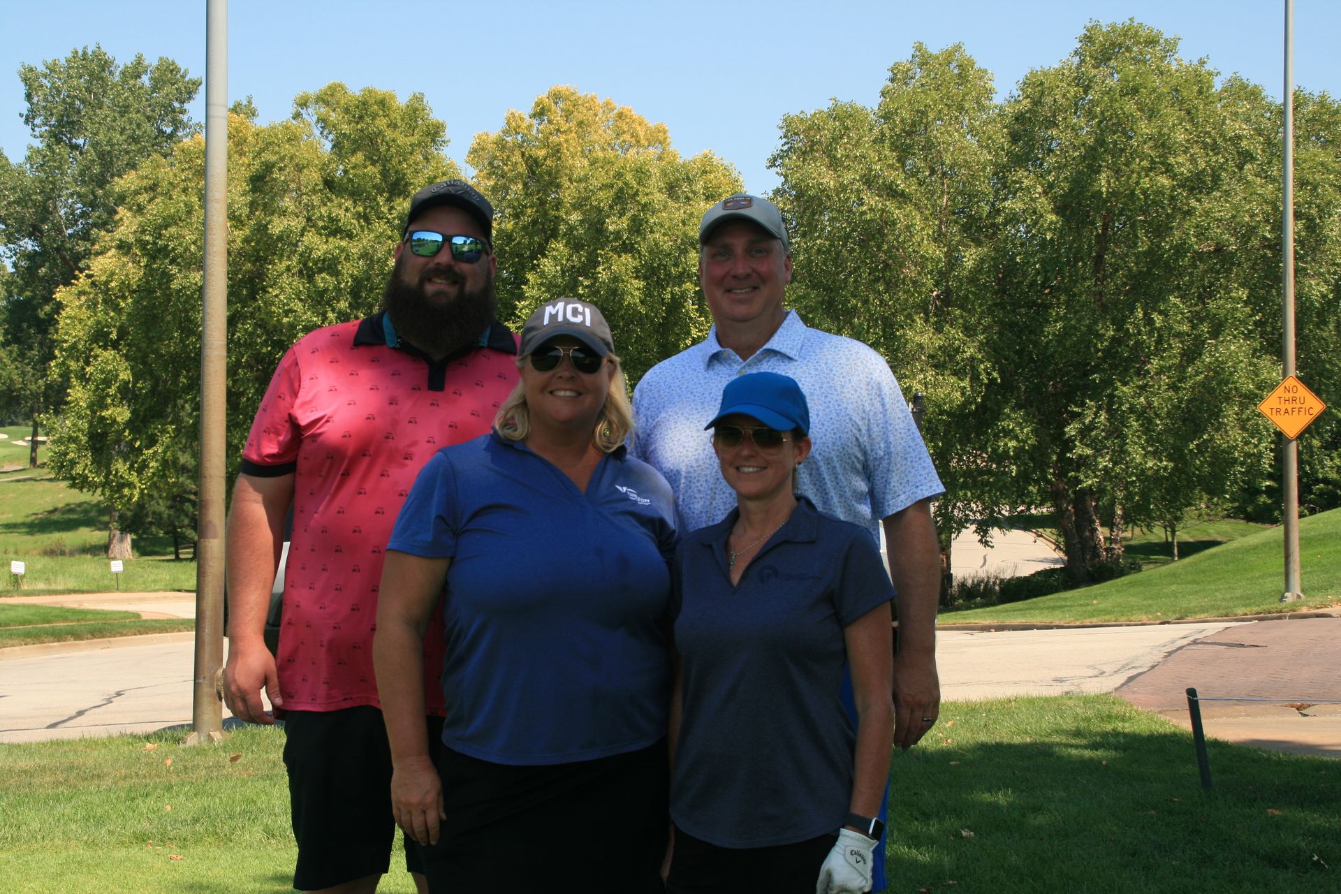 Four people pose on a sunny golf course: a bearded man in pink, two women in blue, and a man in a light blue shirt.