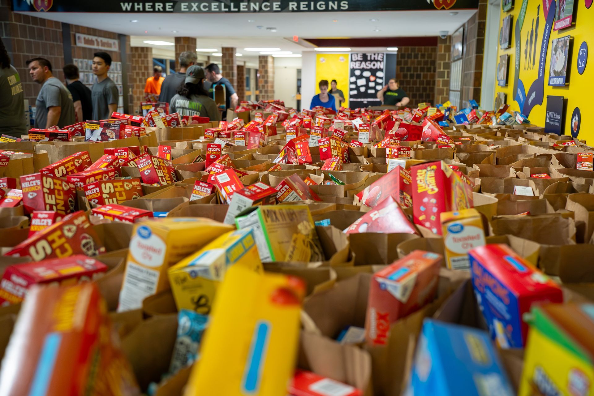 Brown paper bags filled with food, set on tables in a school hallway; people in background.