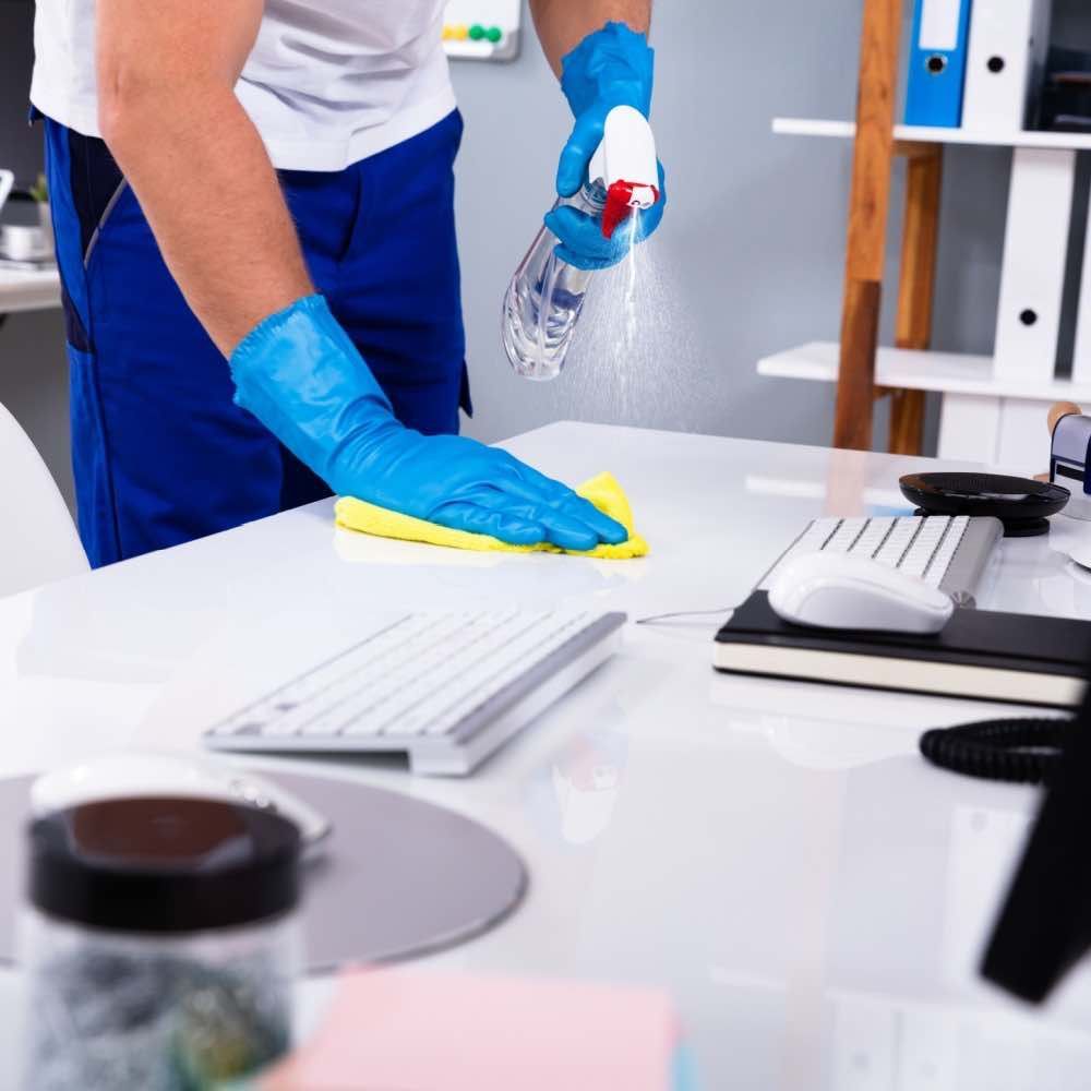 Person in blue gloves spraying and wiping a white desk with cleaning supplies in an office setting.
