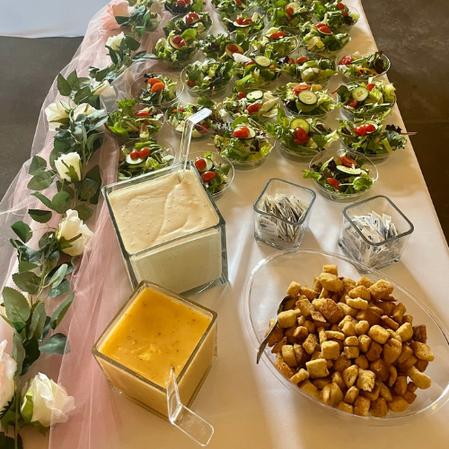 Buffet table with appetizers, salad cups, dipping sauces, and bread cubes on a pink tablecloth