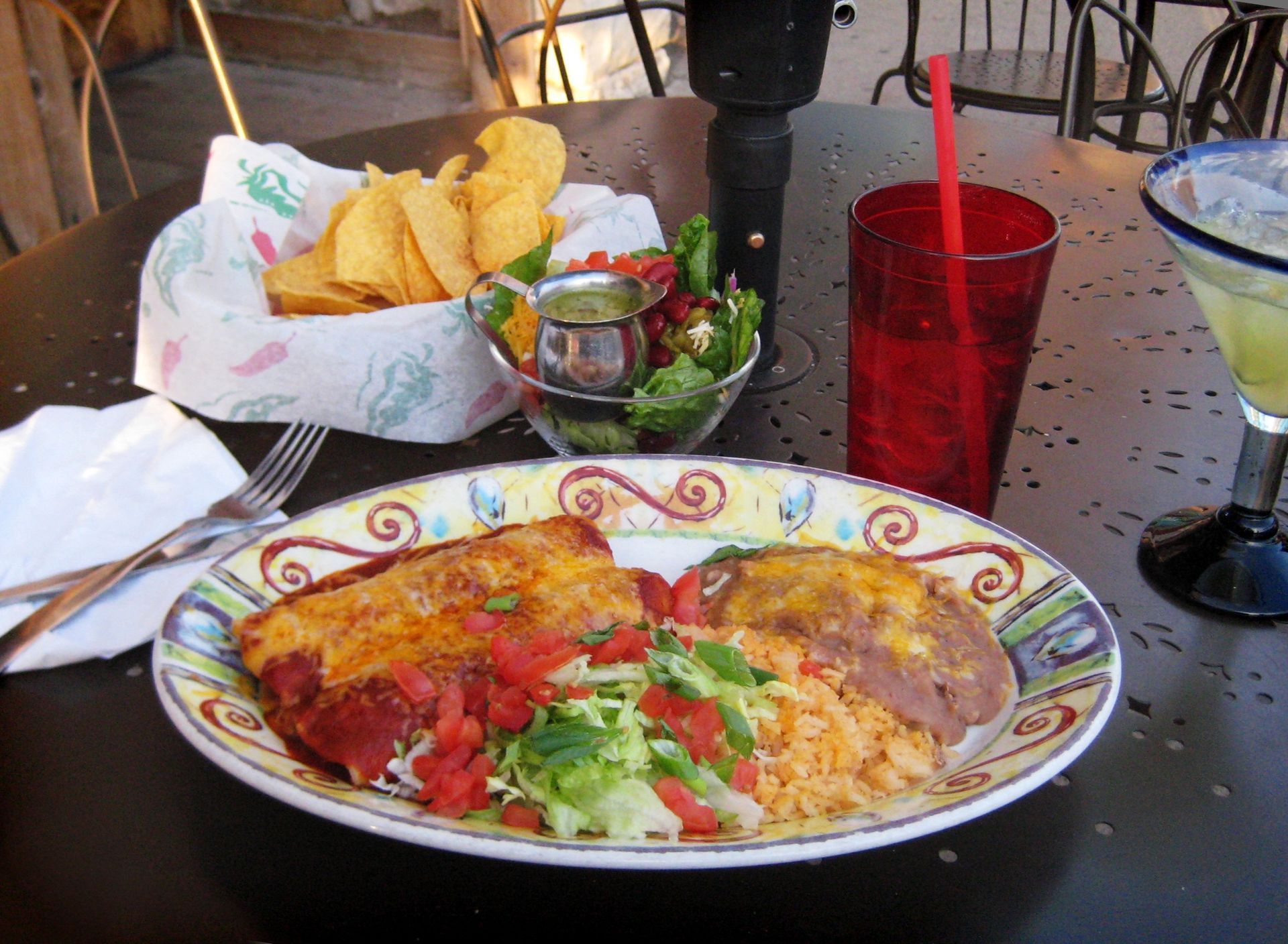 Plated Mexican meal with enchilada, rice, beans, chips, salsa, and a red drink on a table