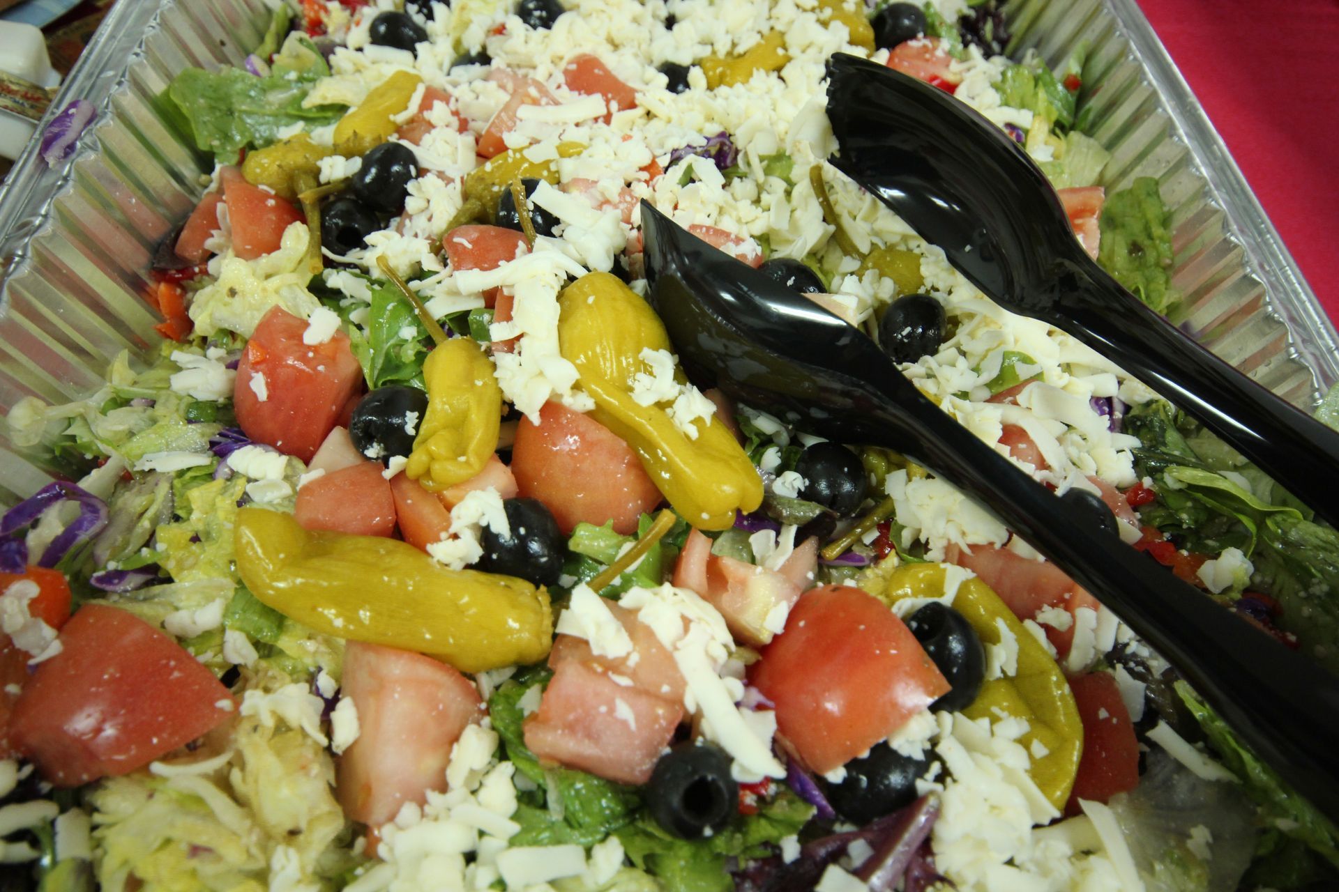 Tray of mixed salad with cherry tomatoes, peppers, shredded cheese, and black serving tongs
