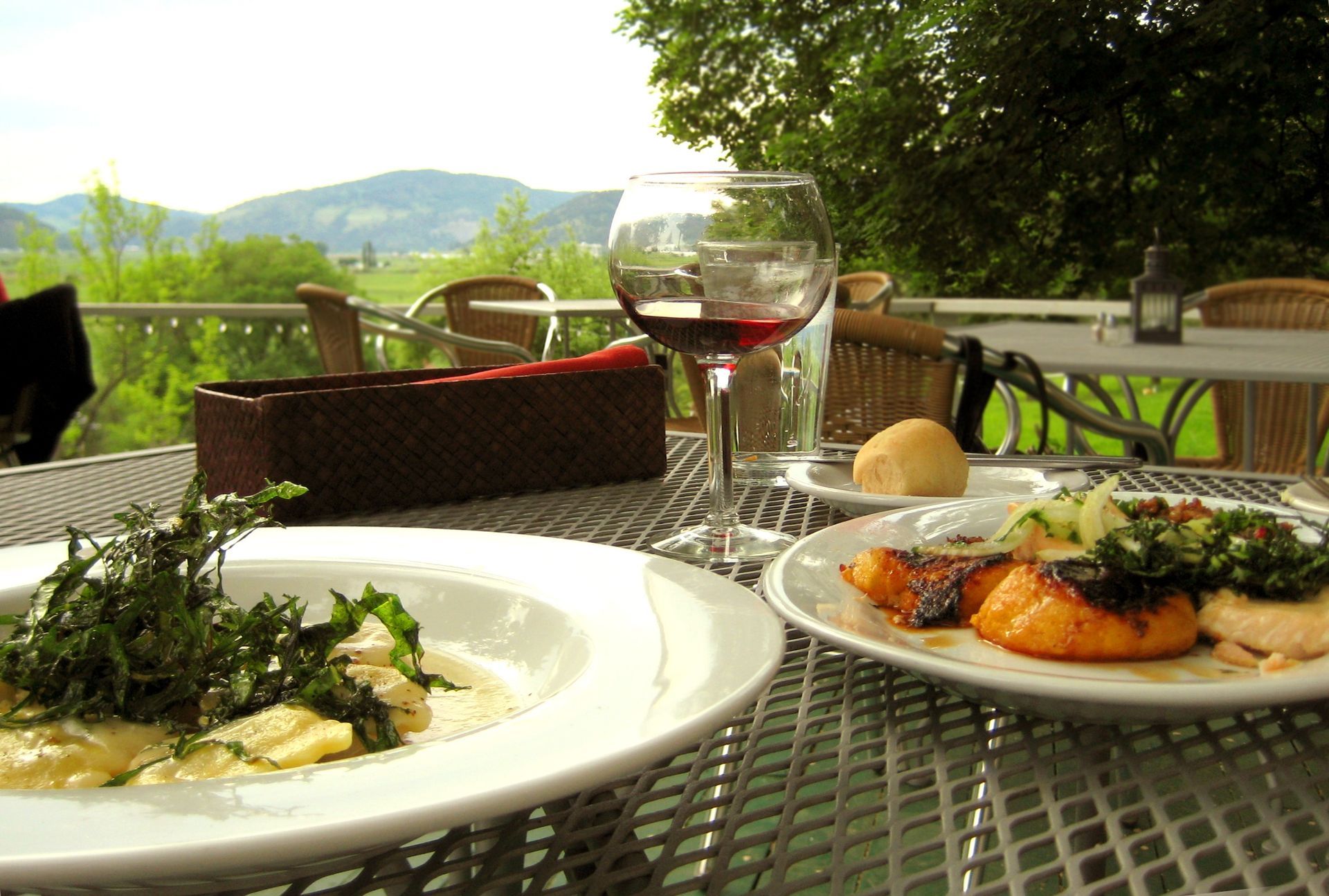 Outdoor patio table with a plated meal, bread, and a glass of red wine overlooking a green hillside