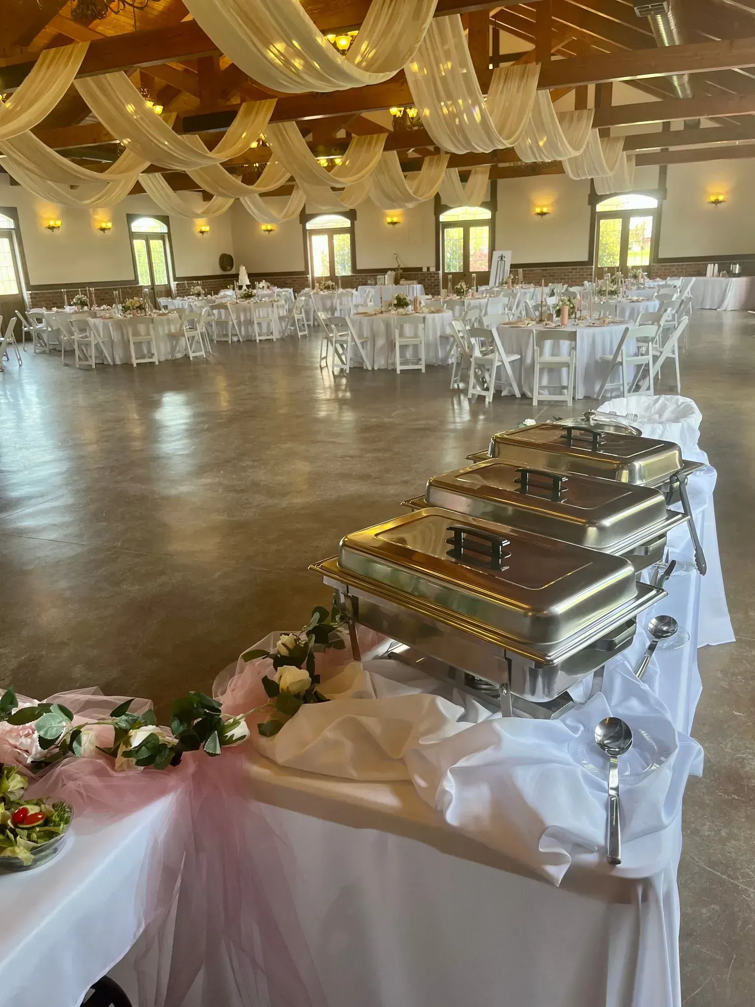 Buffet table with silver chafing dishes in a decorated banquet hall with white chairs and draped gold ceiling.