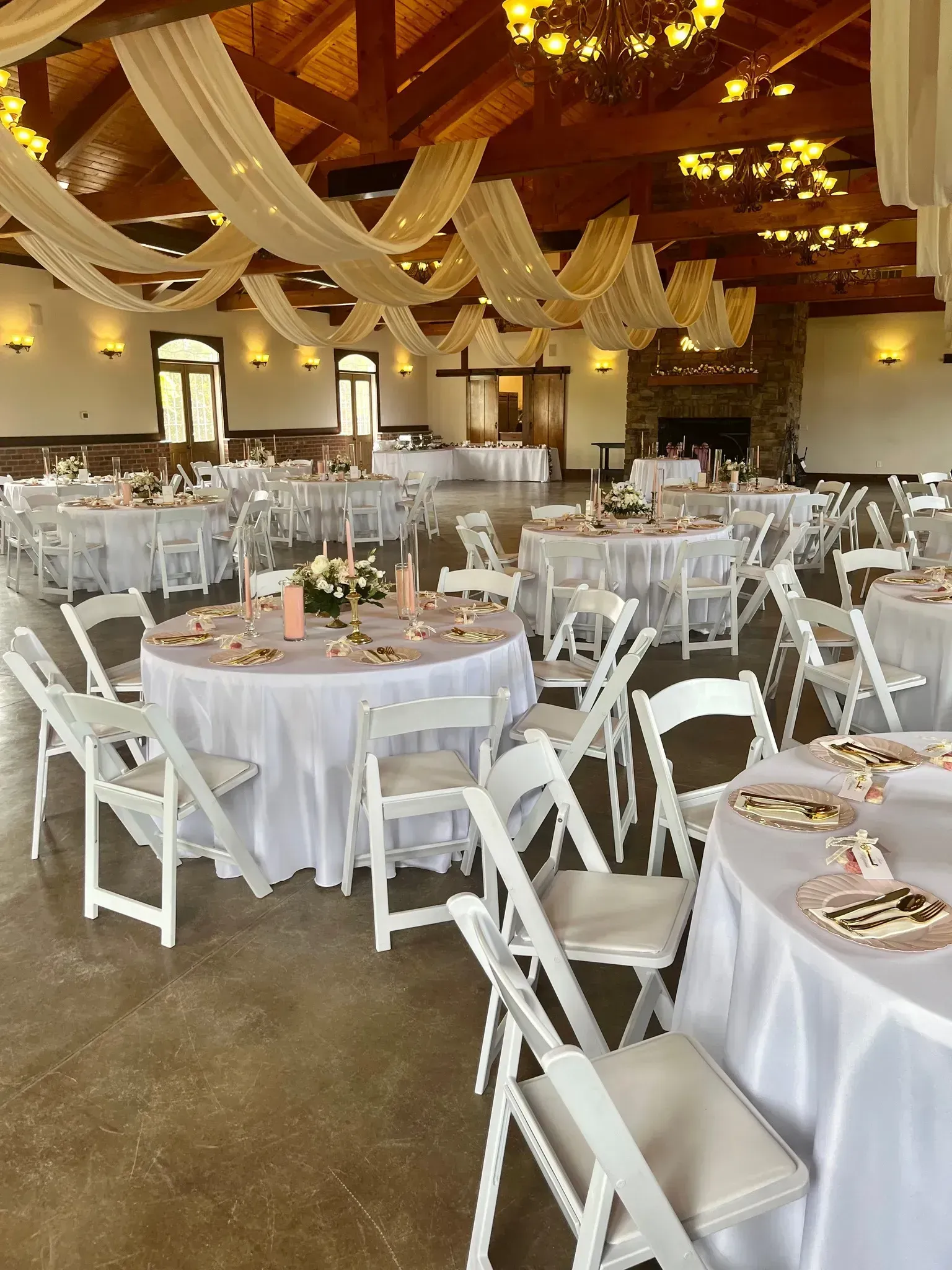 Round banquet tables set for an elegant reception in a warmly lit hall with white chairs and gold ceiling drapes.