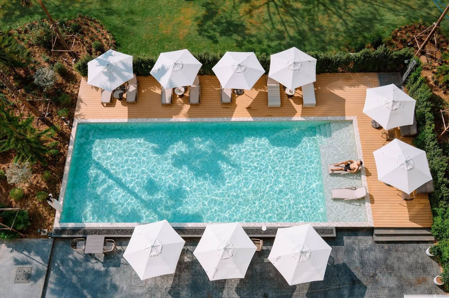 Aerial view of a turquoise pool with white lounge umbrellas and chairs on a wooden deck surrounded by greenery