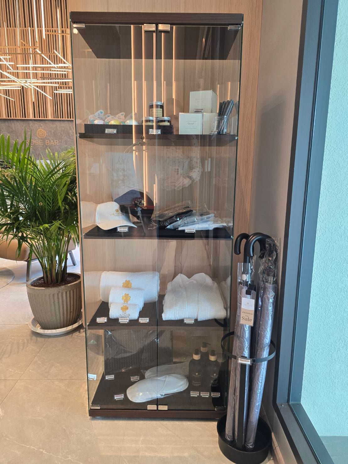 Tall glass display cabinet with neatly folded towels and toiletries beside a plant and water dispenser in a lobby.