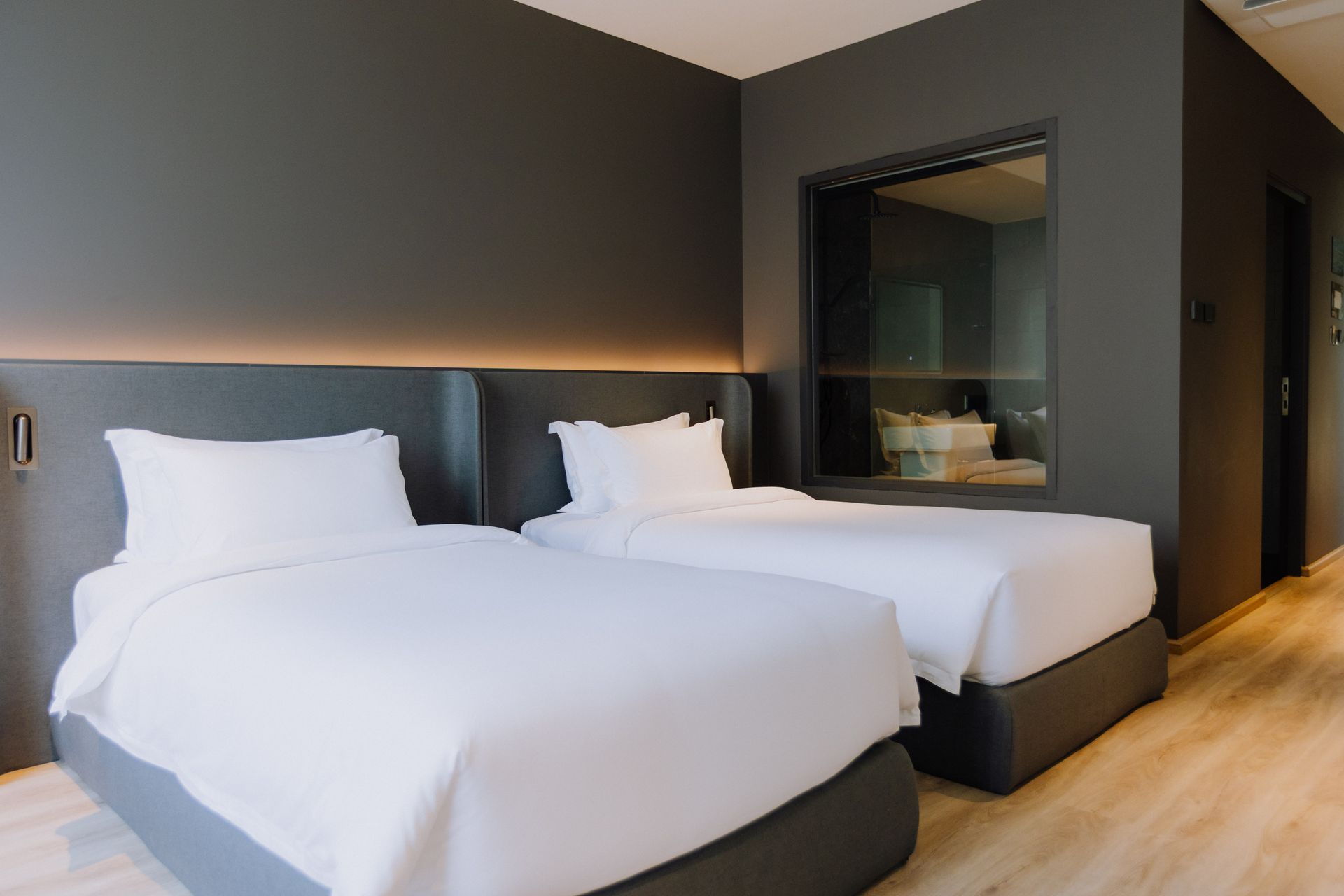 Modern hotel room with two white beds, dark walls, and light wood flooring