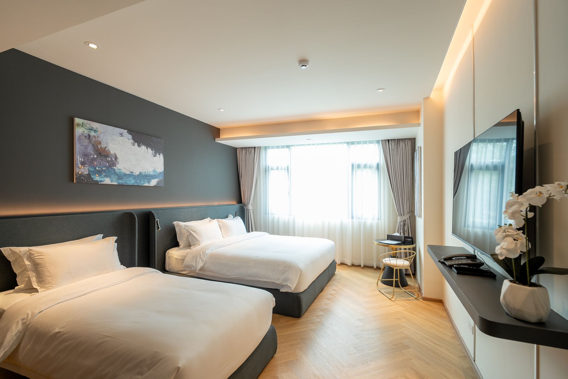 Modern hotel room with two beds, gray accent wall, window seating, and a TV console