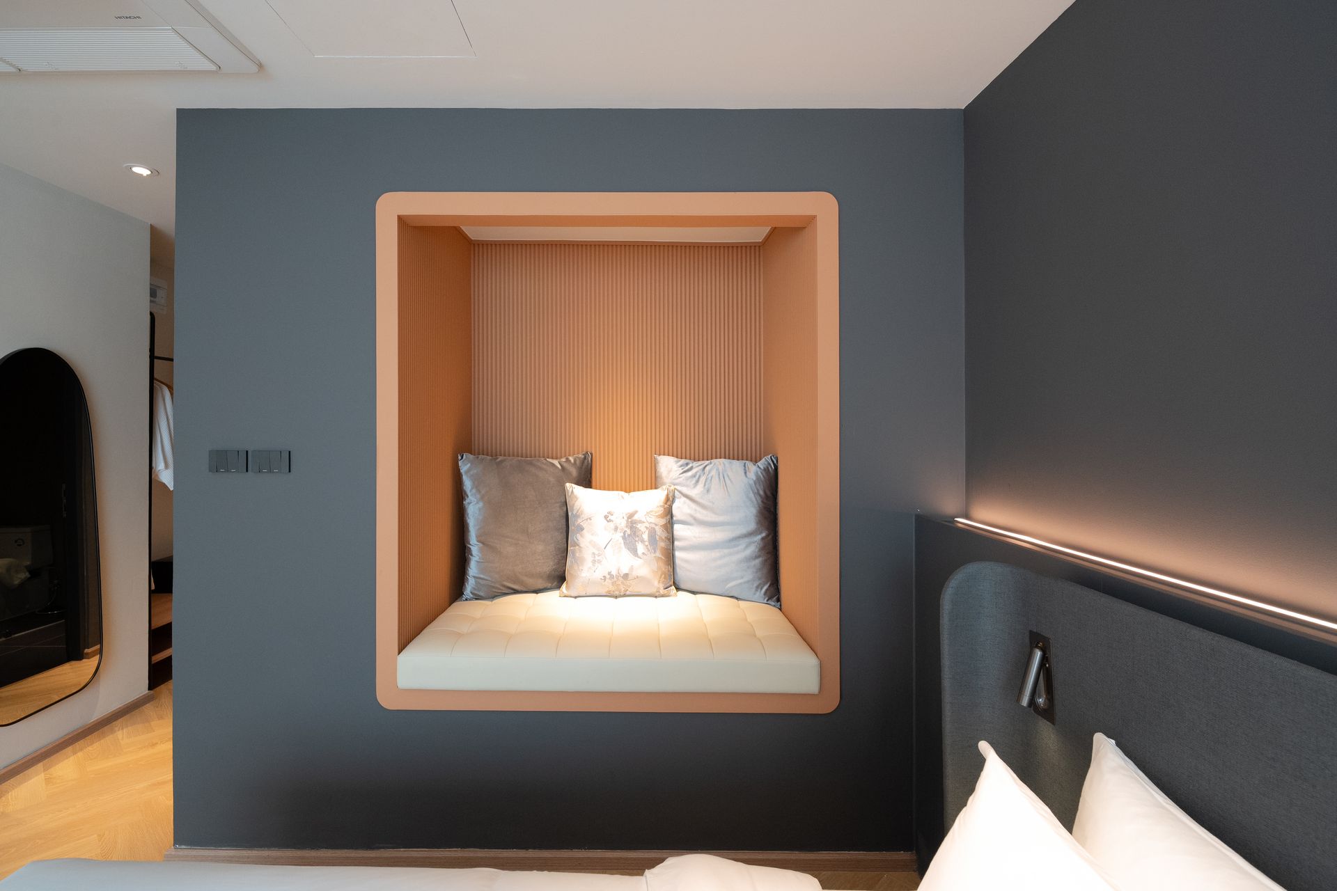 Modern bedroom with a built-in wooden window seat, gray walls, and a bed in the foreground.