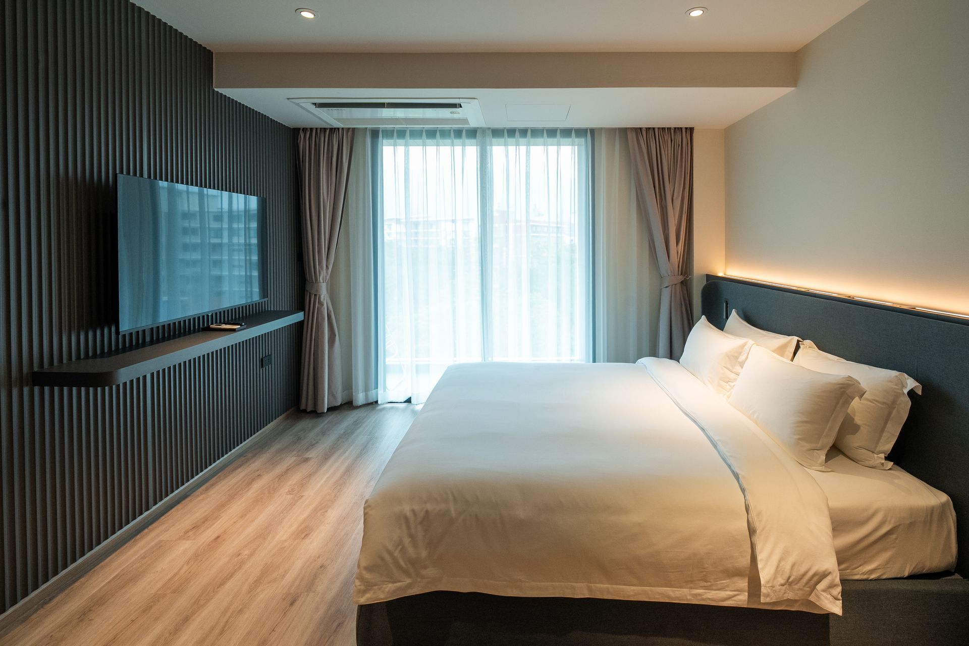 Modern bedroom with a large bed, wall-mounted TV, and sheer curtains letting in daylight