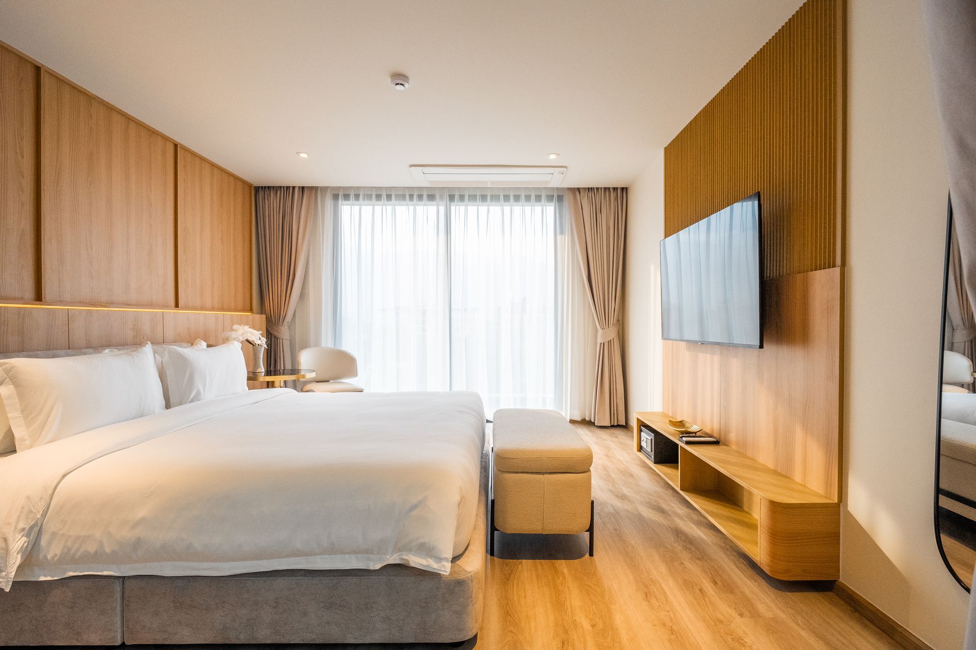 Modern hotel room with a large bed, wood-paneled walls, TV, ottoman, and sheer curtains.