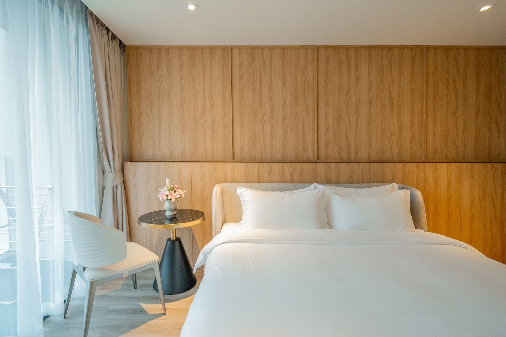 Bright minimalist bedroom with a white bed, wood-paneled wall, and window seating by sheer curtains