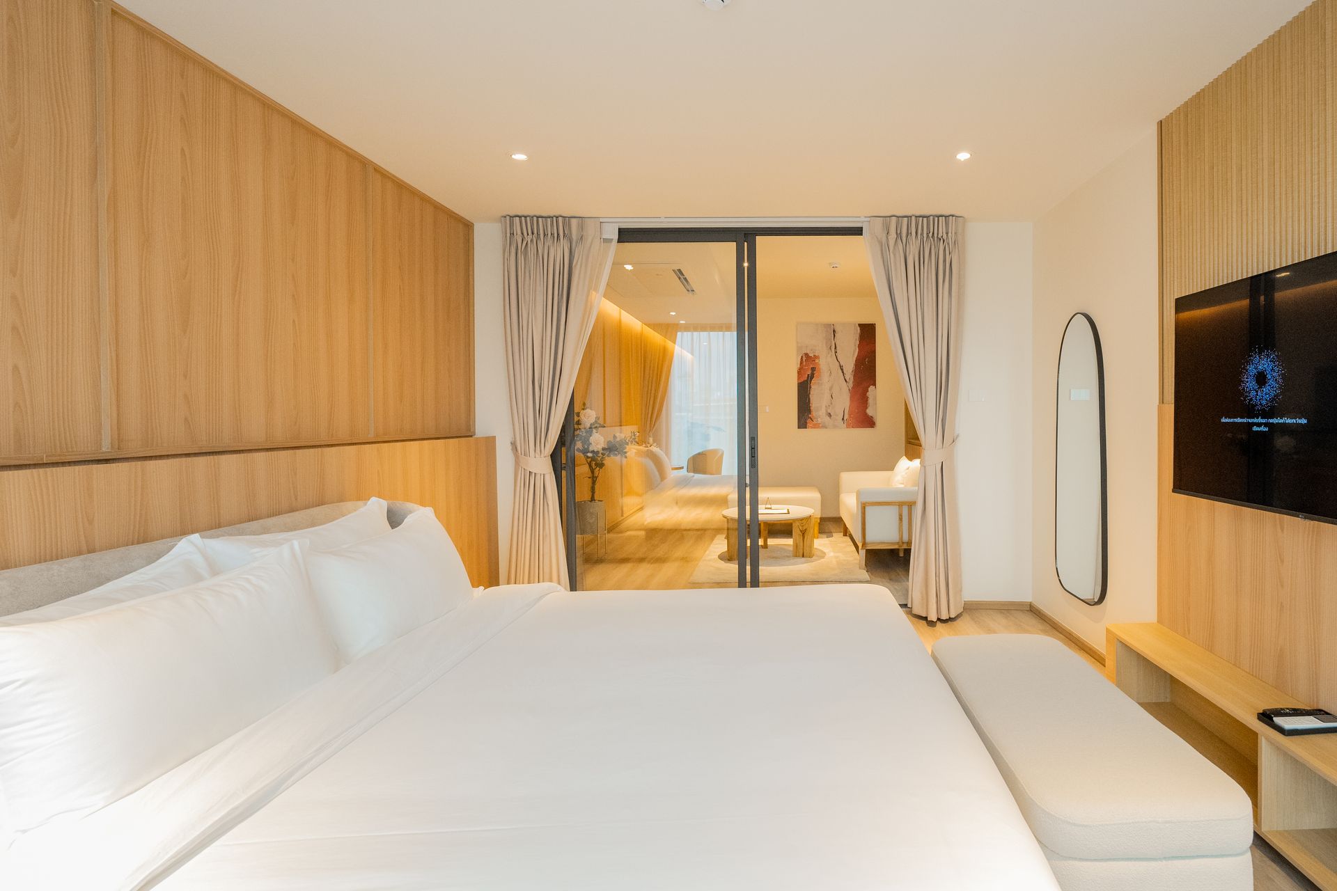 Modern hotel bedroom with a large white bed, wood-paneled walls, and a view into a bright adjacent room