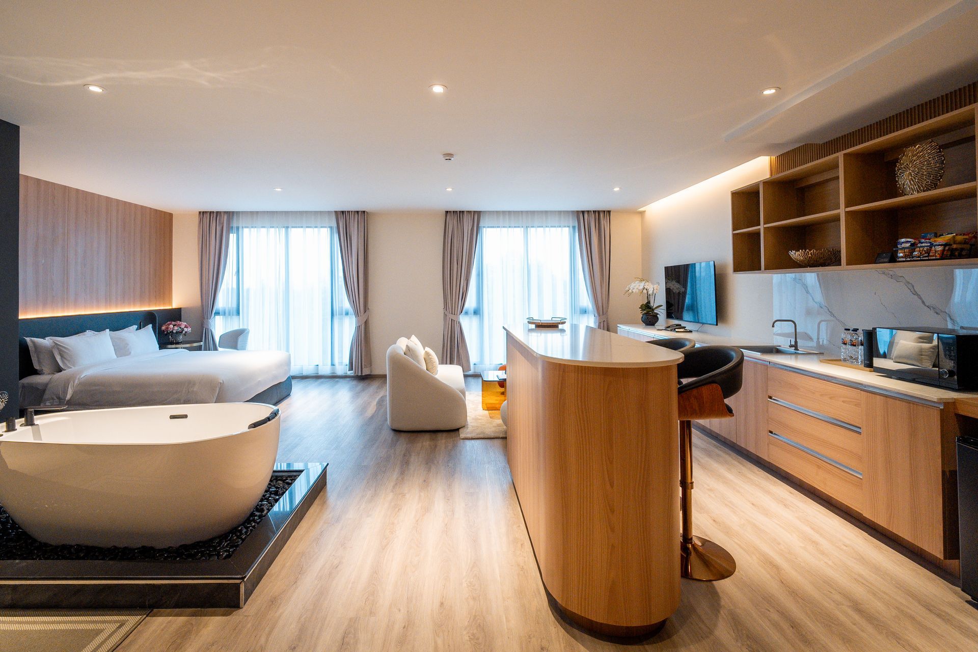Modern hotel room with bed, sofa, desk, and kitchenette in warm wood tones