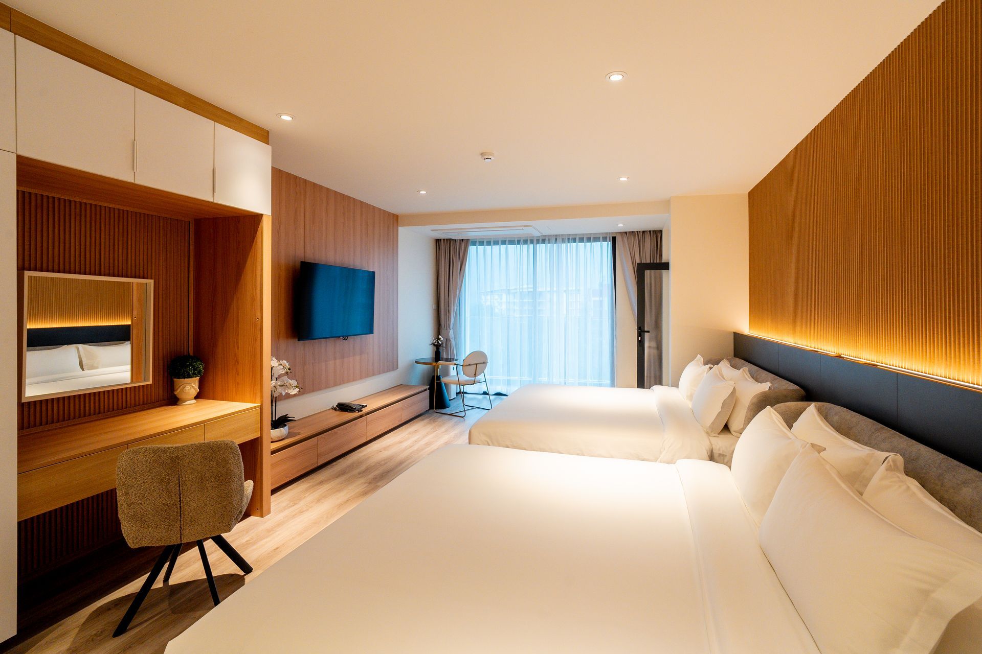 Modern hotel room with two beds, wooden desk, wall-mounted TV, and a large window with sheer curtains