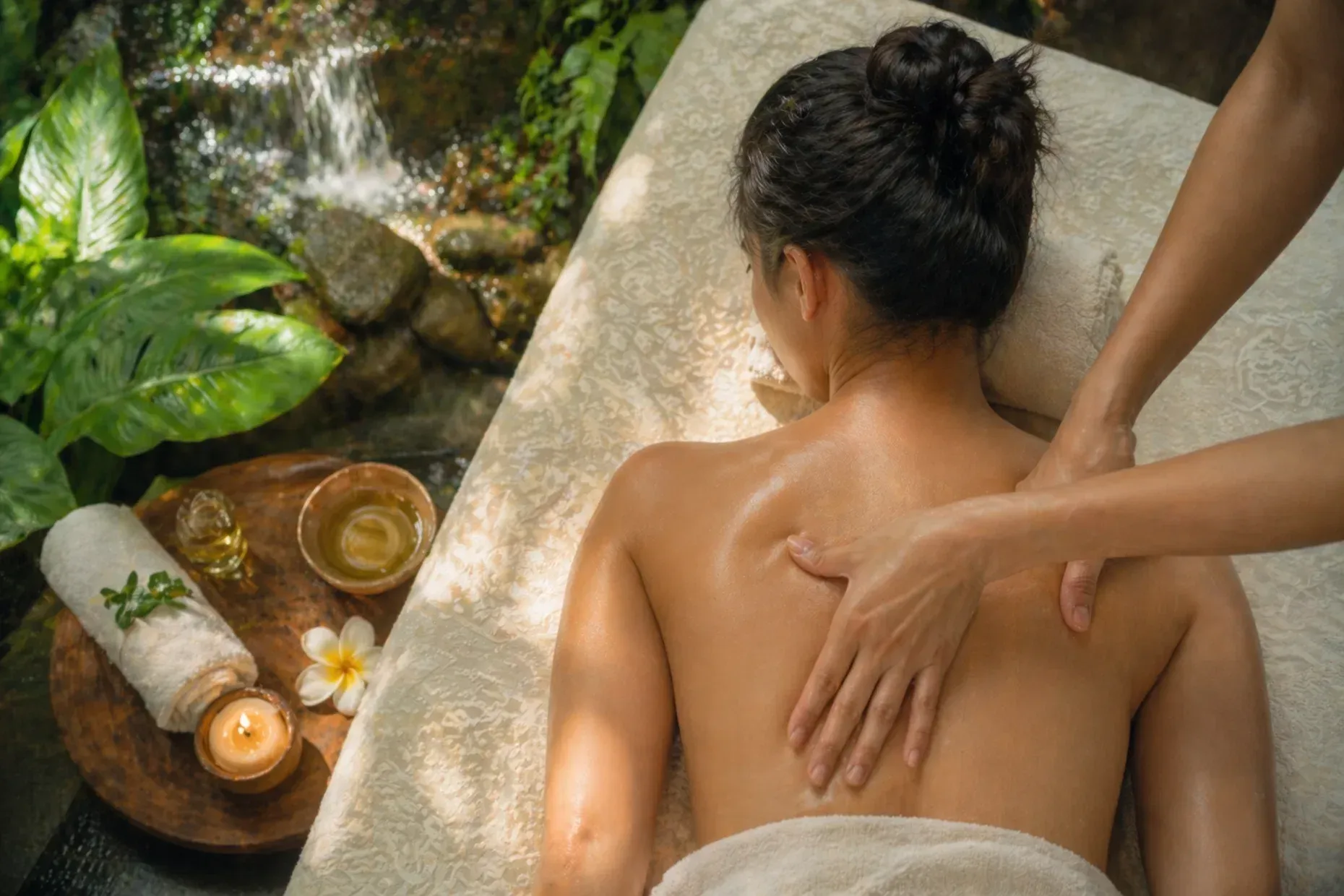 Spa massage by a candlelit waterfall, with an oil bowl and flowers nearby.