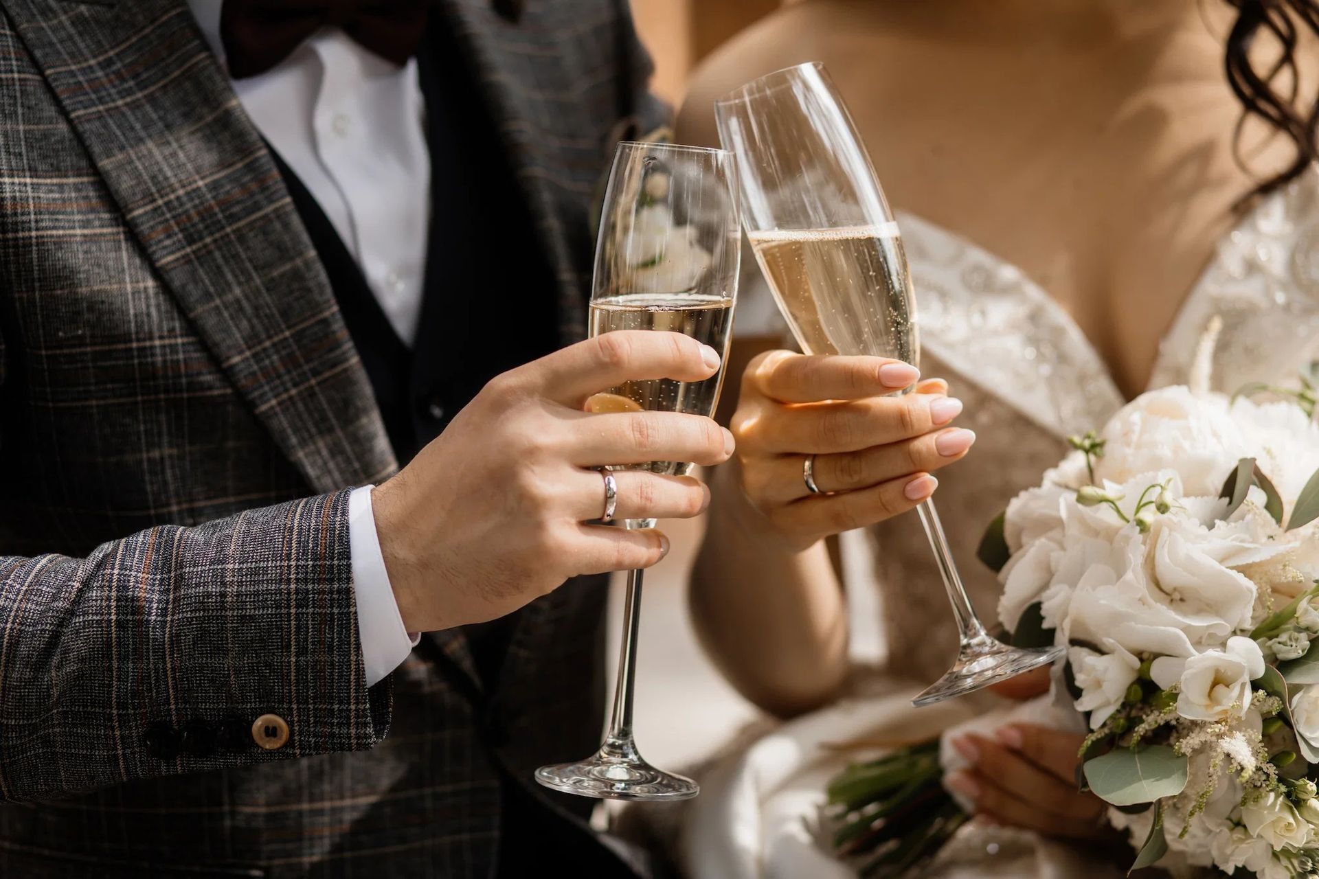 Two people in formalwear toast with champagne, holding a bouquet at a wedding.