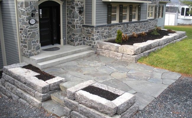 Terrasse en pierre et entrée principale d'une maison avec des marches en béton brut et des murs de soutènement au premier plan.