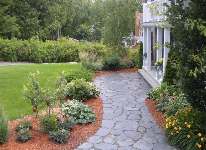 Allée de jardin en dalles de pierre longeant une maison blanche, bordée d'une pelouse verdoyante et de fleurs colorées.