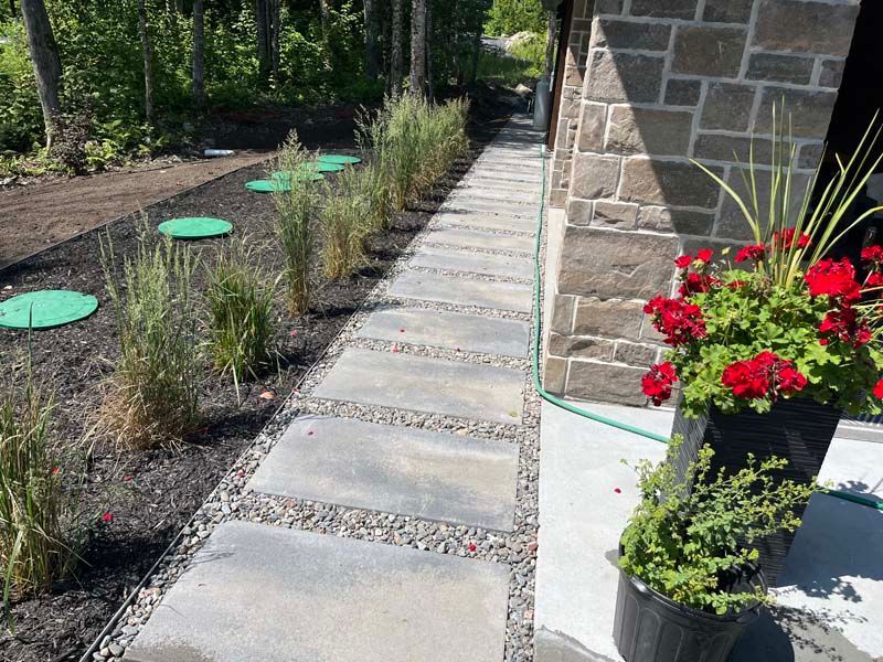 Allée en pierre longeant un parterre de fleurs et un muret en pierre, avec des fleurs rouges en pot près du coin.