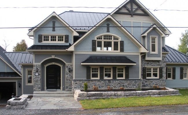 Grande maison grise avec façade en pierre, porche et toit en métal sous un ciel couvert