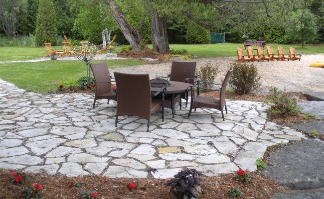 Un coin salon de jardin avec des chaises marron et une table ronde est aménagé sur une terrasse en pierre, à côté d'une pelouse verdoyante et d'arbres.