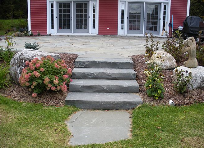 Les marches d'entrée d'une maison rouge aux portes blanches sont bordées de parterres de fleurs et d'une allée en béton.