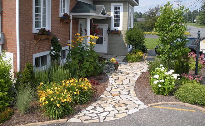Allée en pierre traversant un jardin coloré, à côté d'une maison en briques et d'une allée carrossable.