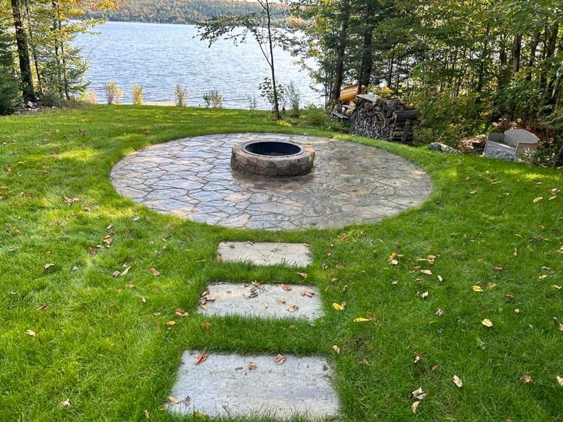 Foyer extérieur en pierre sur une terrasse circulaire donnant sur un lac, avec une pelouse et des arbres.