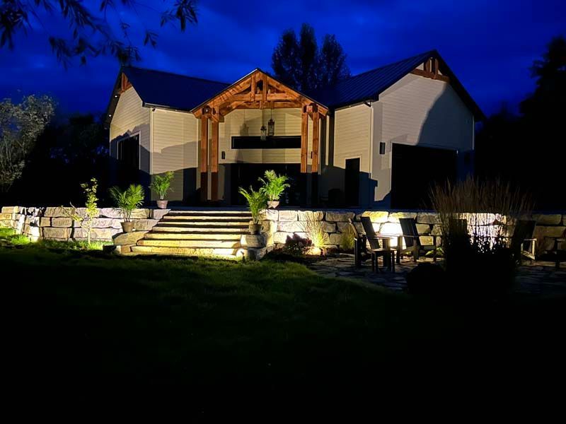 Entrée d'une maison bien éclairée la nuit, avec des marches en pierre, des murs blancs et un porche en bois se détachant sur un ciel bleu foncé.