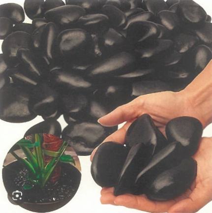 Une main tenant des pierres noires brillantes, avec d'autres pierres noires empilées en arrière-plan et une petite plante en pot en dessous.