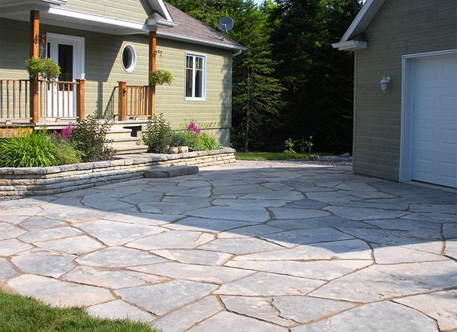 Allée pavée longeant une maison beige avec porche et garage, bordée de parterres de fleurs et d'arbres.