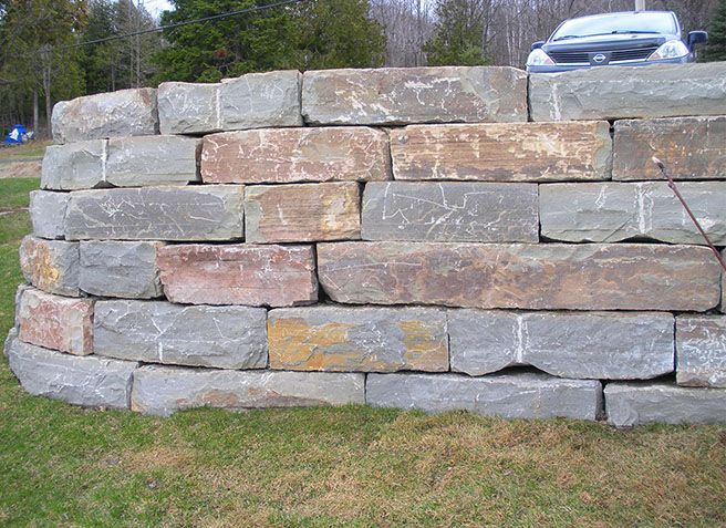 Mur de soutènement en pierres empilées avec une voiture argentée visible au-dessus, dans un espace extérieur herbeux.