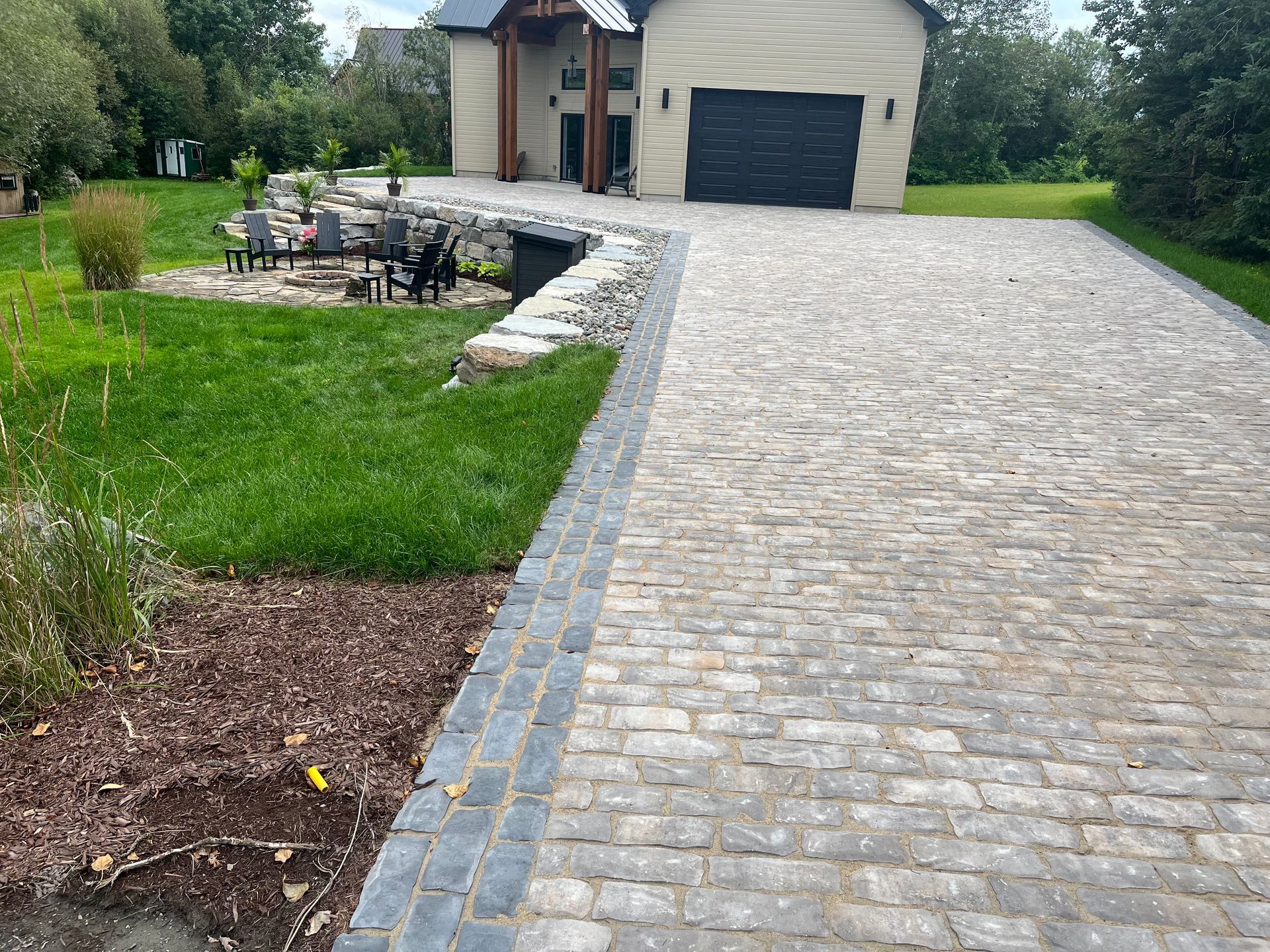Allée pavée menant à une maison beige avec une porte de garage foncée, bordée de pelouse et d'une terrasse.