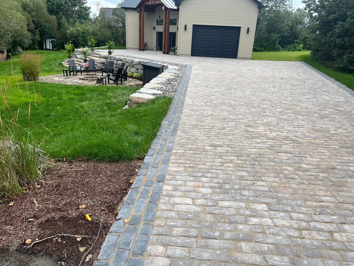 Allée pavée menant à une maison beige avec une porte de garage foncée, bordée de pelouse et d'une terrasse.