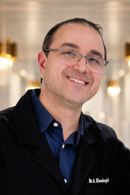 Smiling person in glasses and dark jacket, standing indoors against a softly blurred background.
