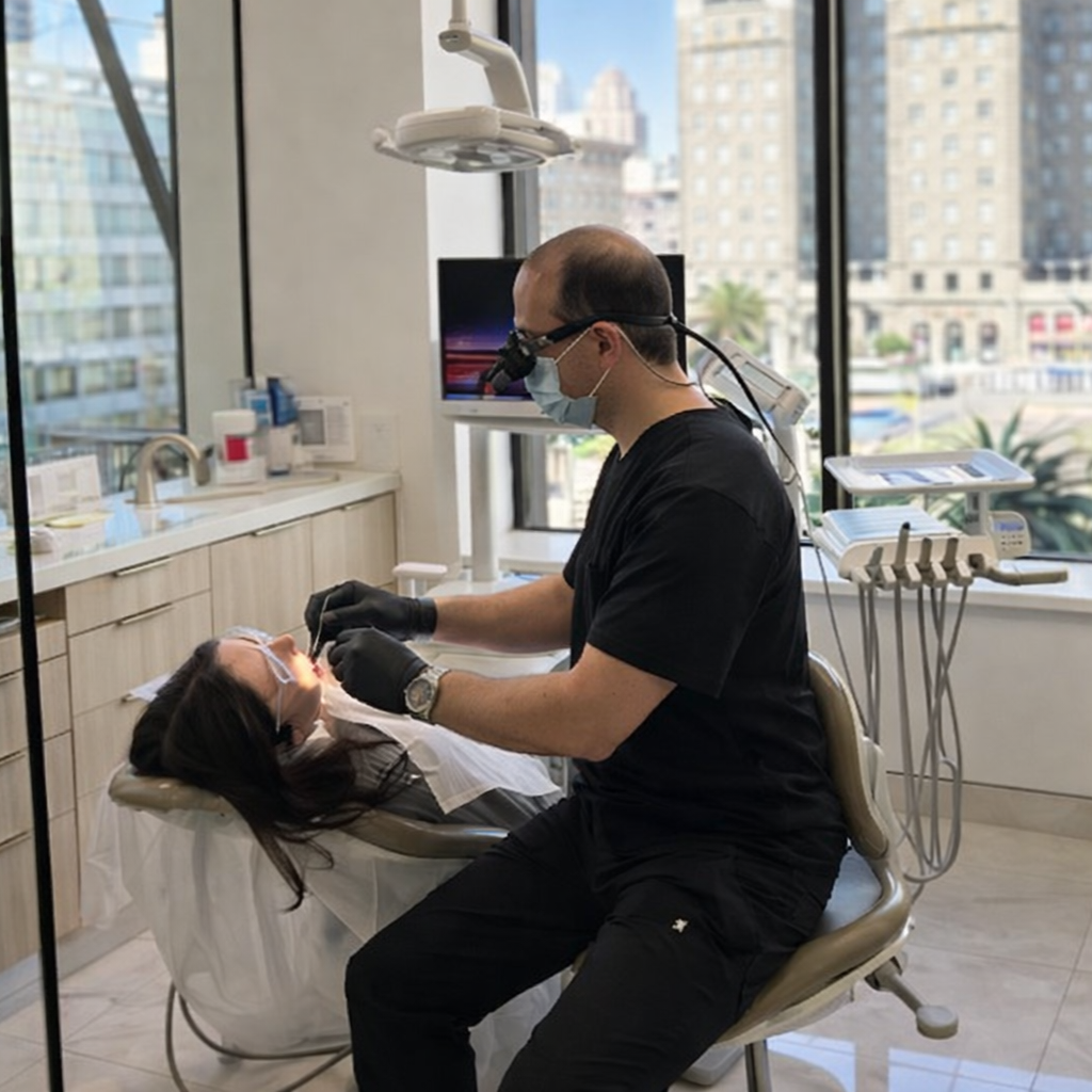 Dentist wearing a mask and gloves treating a patient in a bright dental clinic with city windows.