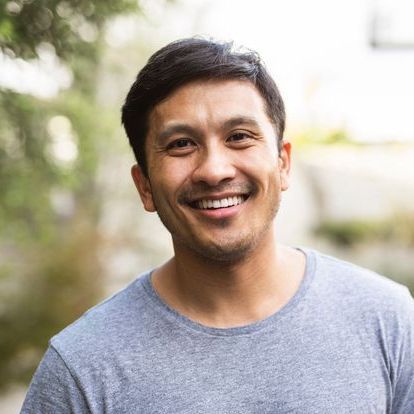 Smiling man in a gray T-shirt standing outdoors with a blurred background