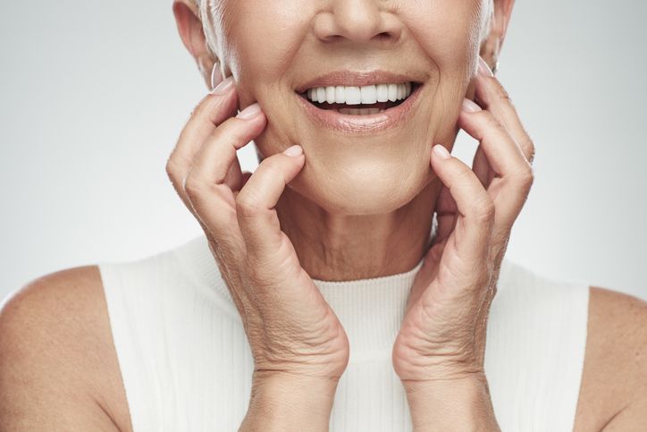 Smiling person touching their cheeks, wearing a white sleeveless top against a light background