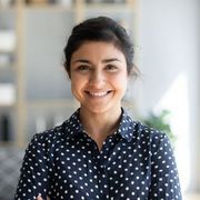 Smiling woman in a navy polka-dot shirt standing indoors against a bright, blurred background