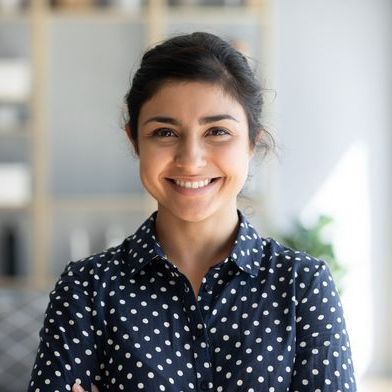 Smiling woman in a navy polka-dot shirt standing indoors against a bright, blurred background