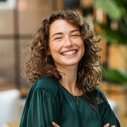 Smiling person with curly hair in a dark green top, standing indoors with a blurred background