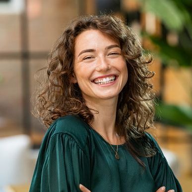 Smiling person with curly hair in a dark green top, standing indoors with a blurred background
